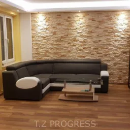 Salon po remoncie: ściana z dekoracyjnej cegły, brązowy narożnik z białymi elementami, drewniana podłoga, ż&oacute;łta ściana, telewizor na szafce RTV.