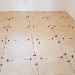 Beżowe płytki podłogowe w trakcie układania, z użyciem czerwono-niebieskich klipsów poziomujących, widoczne linie fug.