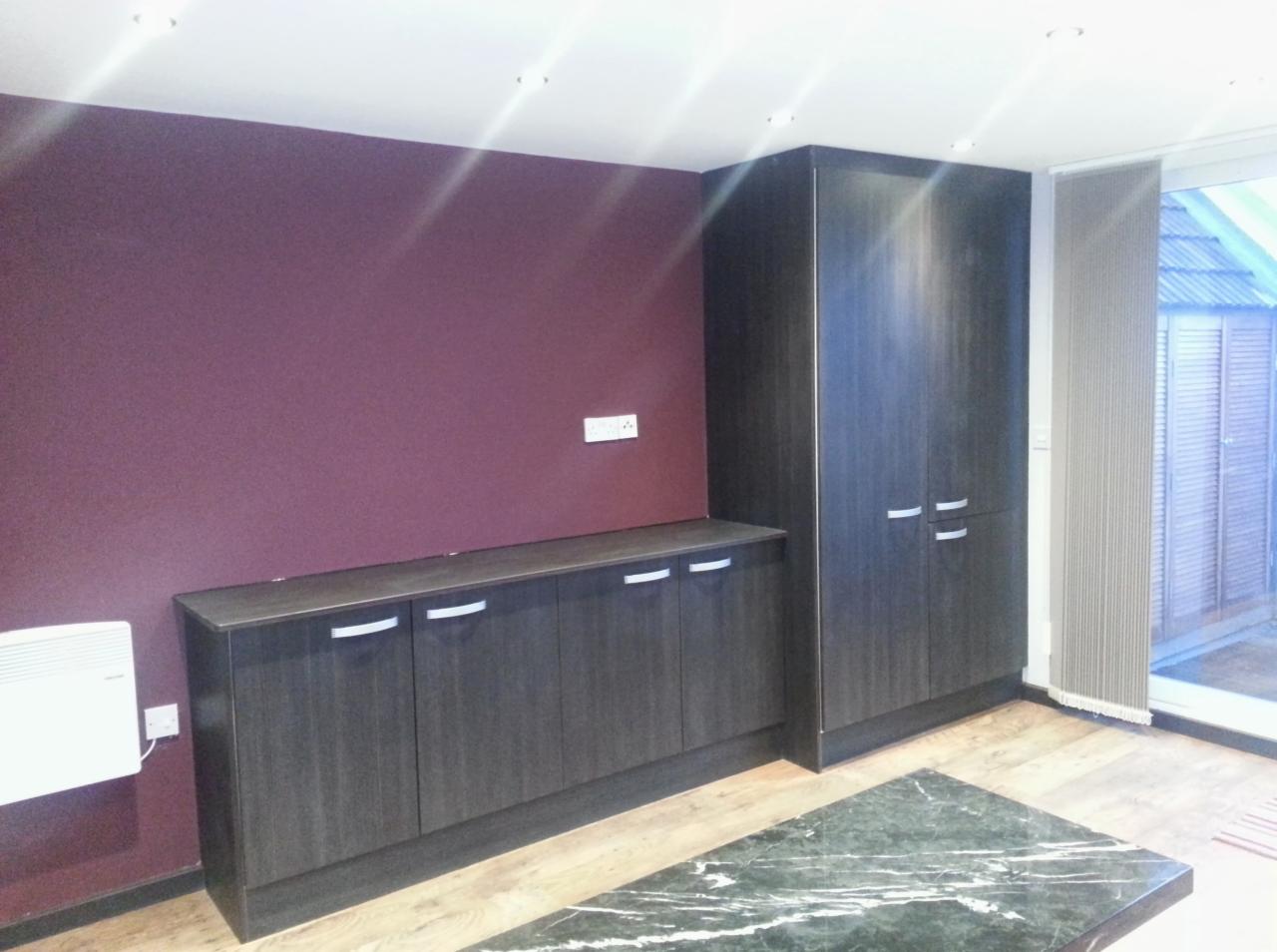 Ściana w kolorze burgundu z zamontowanymi szafkami z ciemnego drewna z poziomymi, srebrnymi uchwytami, biały grzejnik elektryczny i fragment blatu z zielonego marmuru.