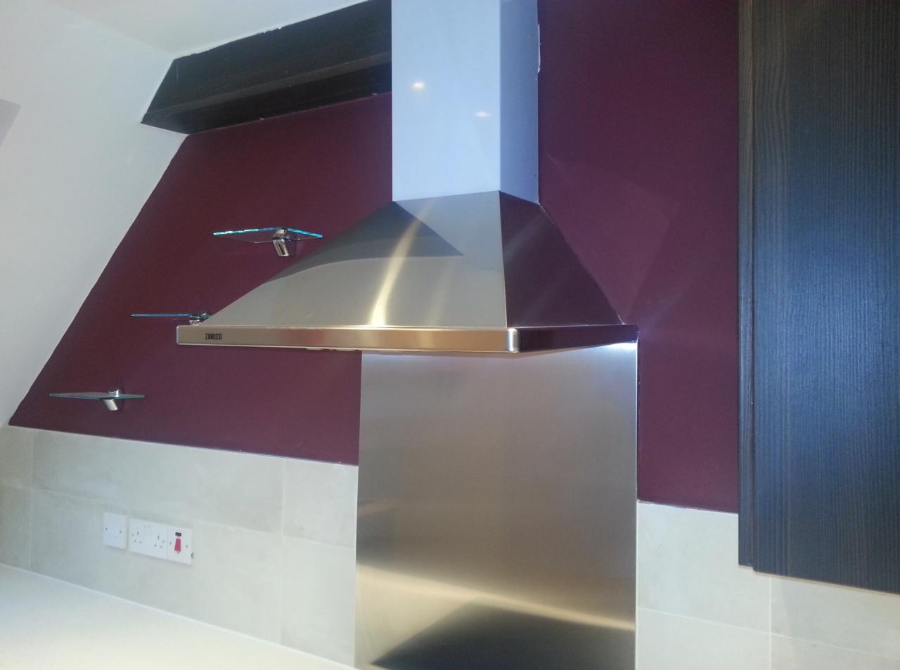 Nowoczesny okap kuchenny ze stali nierdzewnej, zamontowany na tle ściany pomalowanej na ciemny fiolet, z trzema szklanymi półkami po lewej stronie i ciemną szafką po prawej.