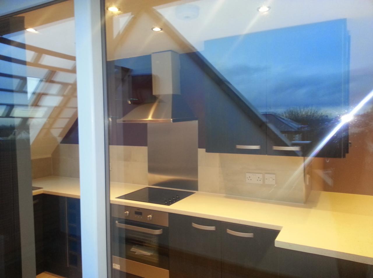 Nowoczesna kuchnia z ciemnymi szafkami, jasnym blatem i stalowym okapem, widoczna przez szybę z odbiciem nieba i dachu sąsiedniego budynku.