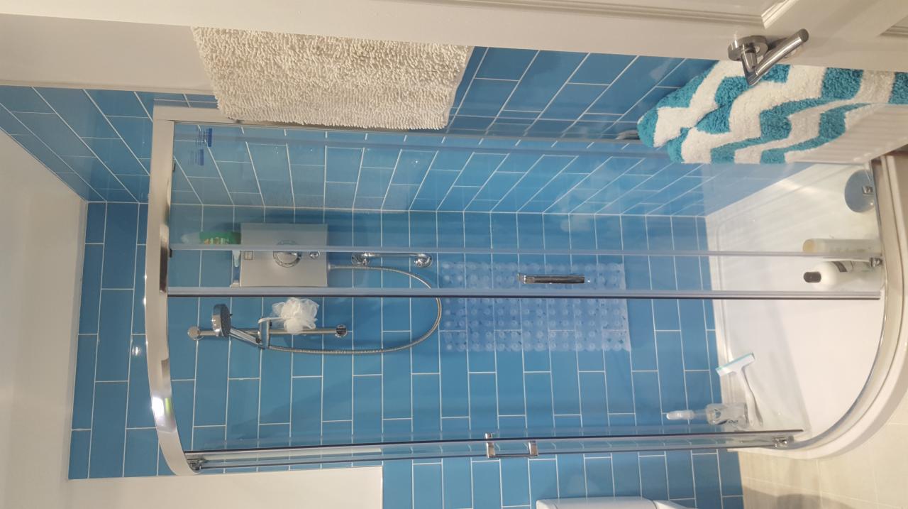 Niebieskie, pionowo ułożone płytki w kabinie prysznicowej z przesuwanymi drzwiami w Trzydniku Dużym. Widoczny biały dywanik i ręcznik z wzorem chevron.