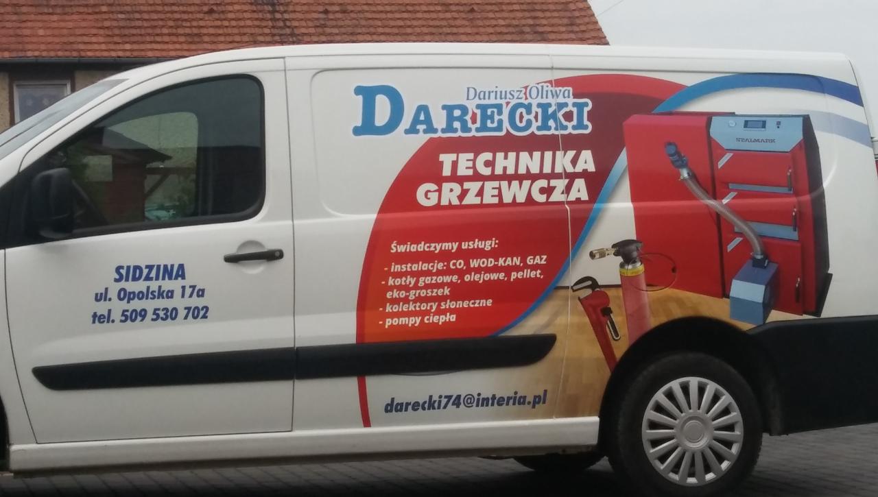 Biały samochód dostawczy z reklamą firmy Darecki Technika Grzewcza, prezentujący grafikę kotła grzewczego, adres firmy w Sidzinie i numer telefonu.
