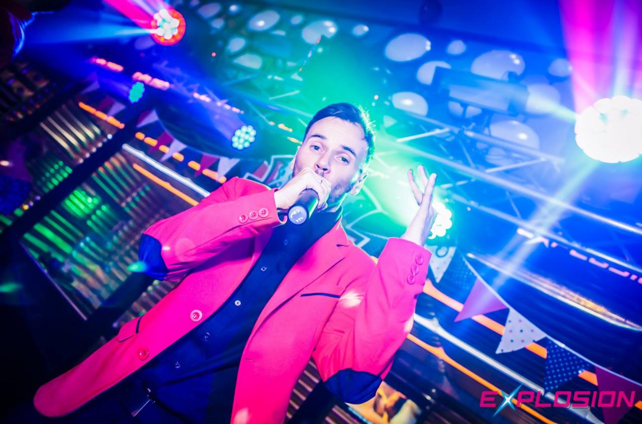 Wokalista w różowej marynarce śpiewa do mikrofonu na scenie oświetlonej kolorowymi światłami, z dekoracjami w tle.