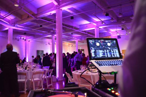 DJ sprzęt (laptop i mikser) na tle sali bankietowej z gośćmi, oświetlonej fioletowym światłem.