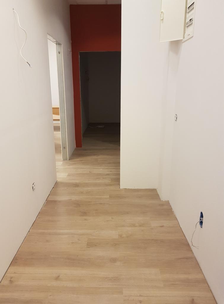 Korytarz w trakcie remontu: świeżo położona podłoga z paneli, białe ściany w trakcie malowania, widoczne puszki elektryczne bez osprzętu, przejście do pokoju z pomalowaną na czerwono ścianą.