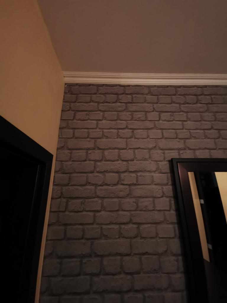 Fragment ściany z tapetą imitującą cegłę w odcieniach szarości, zakończonej białym dekoracyjnym gzymsem pod sufitem, widoczny fragment czarnej framugi drzwi i ramy lustra.