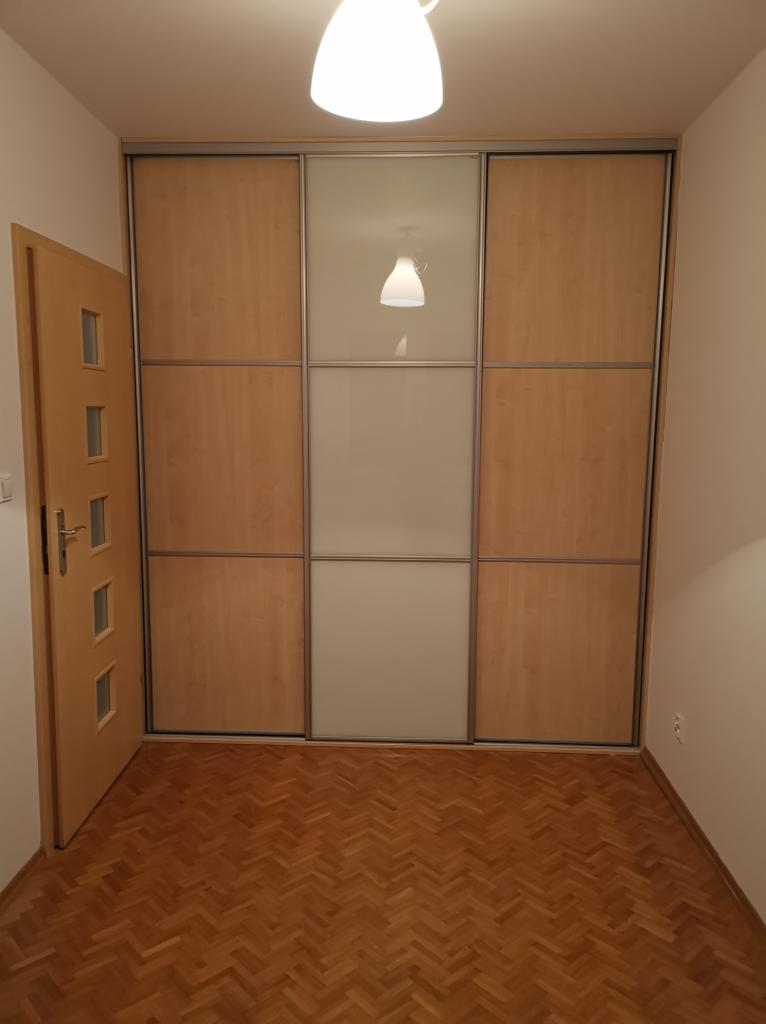 Wnętrze przedpokoju z drewnianą podłogą w jodełkę i zabudowaną szafą przesuwną z panelami drewnianymi i mlecznym szkłem, oświetlone lampą sufitową.