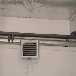 Nagrzewnica wodna Volcano EC zamontowana na ścianie, z widocznymi rurami instalacji grzewczej i izolacją sufitu.