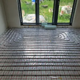 Instalacja ogrzewania podłogowego z rurkami grzewczymi ułożonymi na folii z czerwoną siatką, widok z góry w pomieszczeniu z oknem wychodzącym na zewnątrz.