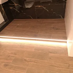 Nowoczesna łazienka z podświetlaną podłogą z płytek imitujących drewno i czarnymi, marmurkowymi płytkami na ścianie.