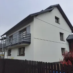 Dwupiętrowy dom z białymi ścianami po renowacji w Proszowicach, widoczny zza ciemnego drewnianego płotu, z metalowymi rusztowaniami przy balkonie.