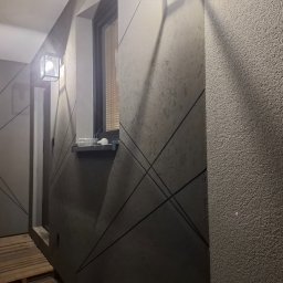 Adam Bawoł Firma Usługowo-Budowlana - Szara ściana z dekoracyjną fakturą i geometrycznym wzorem z ciemnych linii. Widoczne okno z żaluzjami i lampa ścienna. Częściowo widoczna podłoga z drewnianych desek.