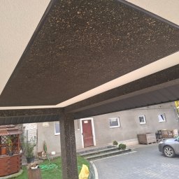 Adam Bawoł Firma Usługowo-Budowlana - Zadaszenie tarasu z dekoracyjną, ciemną podbitką w kształcie trapezu, otoczone jasnym sufitem. Widoczny fragment domu i altana w tle.