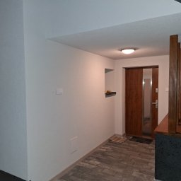 Adam Bawoł Firma Usługowo-Budowlana - Wykończony korytarz z drewnianymi drzwiami, szarą podłogą z płytek imitujących drewno i świeżo pomalowanymi ścianami w odcieniach bieli i szarości. Widoczny fragment instalacji.