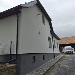 Usługi budowlane i elewacyjne - Dwukolorowa elewacja domu z szarą podmurówką i białymi ścianami. Widoczny fragment dachu, rynny i podjazd z kostki. W tle budynek gospodarczy i samochód dostawczy.