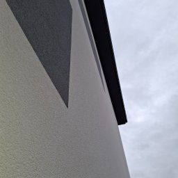 Adam Bawoł Firma Usługowo-Budowlana - Fragment elewacji budynku z dekoracyjnym, geometrycznym wzorem w odcieniach szarości i beżu, widoczne orynnowanie na tle pochmurnego nieba.
