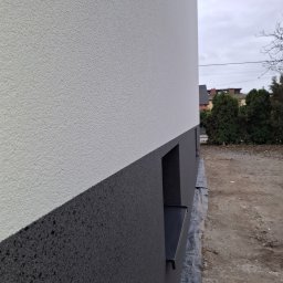 Adam Bawoł Firma Usługowo-Budowlana - Dwukolorowa elewacja budynku z wyraźną fakturą tynku, widok z boku, z fragmentem okna i zabezpieczeniem gruntu folią. Szara, pochmurna pogoda w tle.