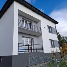 Dwupiętrowy dom z balkonami, świeżo po remoncie elewacji w odcieniach szarości i bieli, z czarnymi balustradami. Widok z dołu, błękitne niebo w tle.