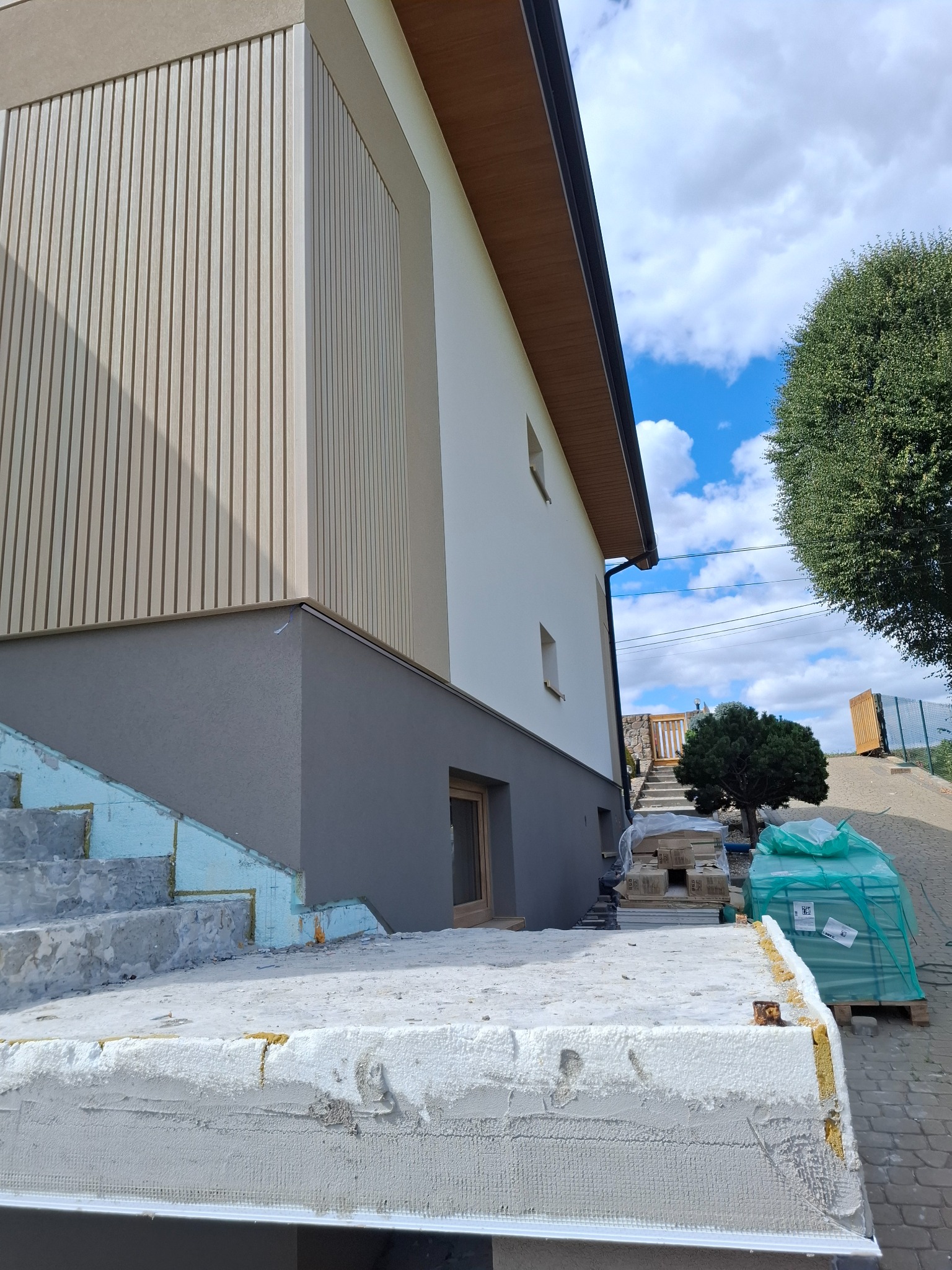 Nowoczesna elewacja domu z drewnianymi panelami i szarą podmurówką. Widoczne ocieplone schody wejściowe i materiały budowlane. Błękitne niebo z chmurami w tle.