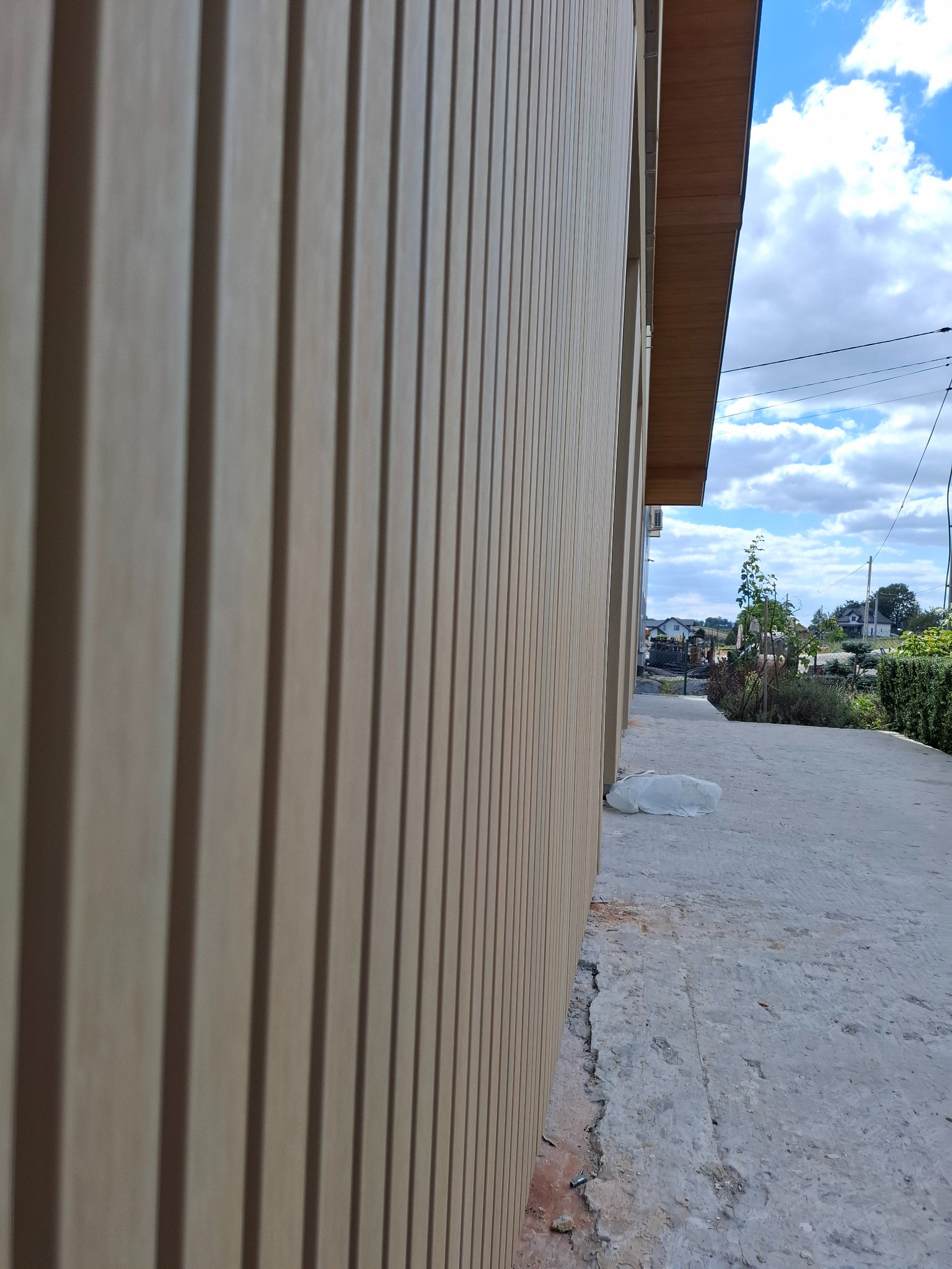 Beżowa elewacja budynku z pionowymi panelami, widok z boku, z fragmentem chodnika i niebem w tle. Nowoczesne wykończenie zewnętrzne.