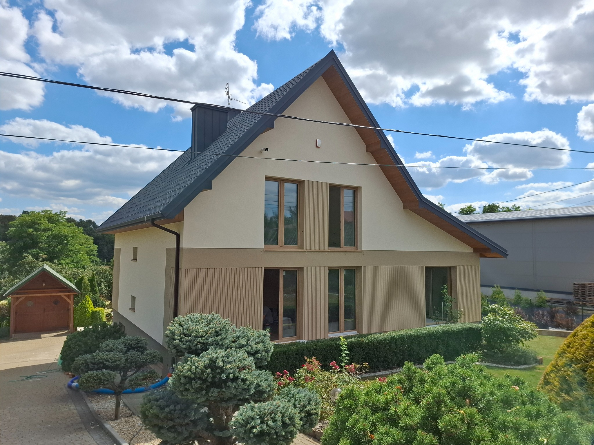 Dwukondygnacyjny dom z beżową elewacją i drewnianymi akcentami, otoczony zielenią. Widoczne okna i ciemny dach. Błękitne niebo z chmurami w tle.