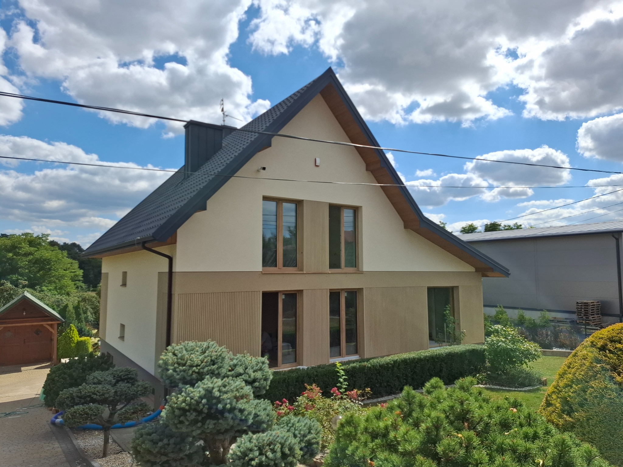 Dwukondygnacyjny dom z beżową elewacją i ciemnym dachem na tle błękitnego nieba z chmurami. Widoczne drewniane okna i zadbany ogród z krzewami.