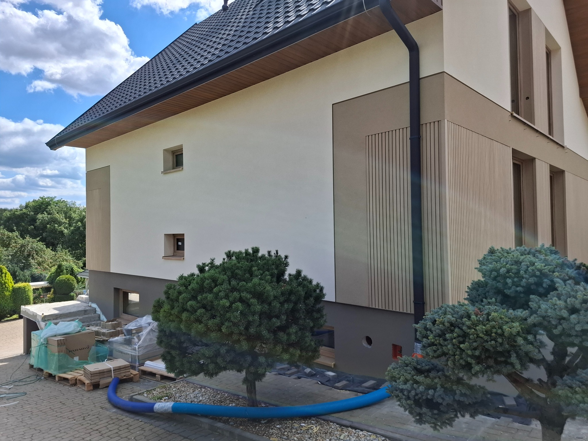 Nowoczesny dom z elewacją w odcieniach bieli i beżu, z elementami dekoracyjnymi z pionowych paneli. Widoczne materiały budowlane i niebieski wąż na posesji.