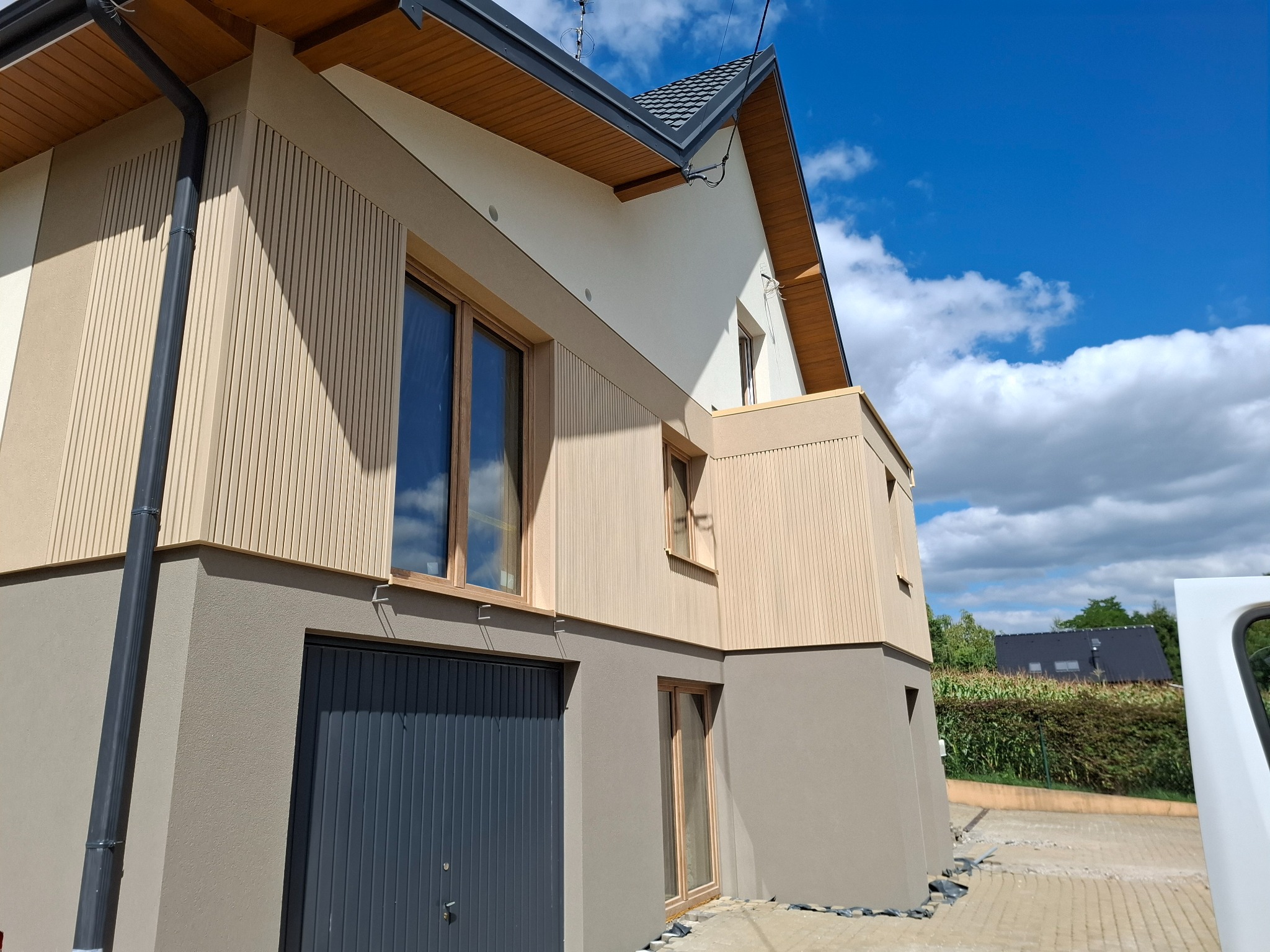 Dwukondygnacyjny dom z elewacją w odcieniach beżu i szarości, z drewnianymi akcentami i ciemną stolarką okienną, na tle błękitnego nieba z chmurami.