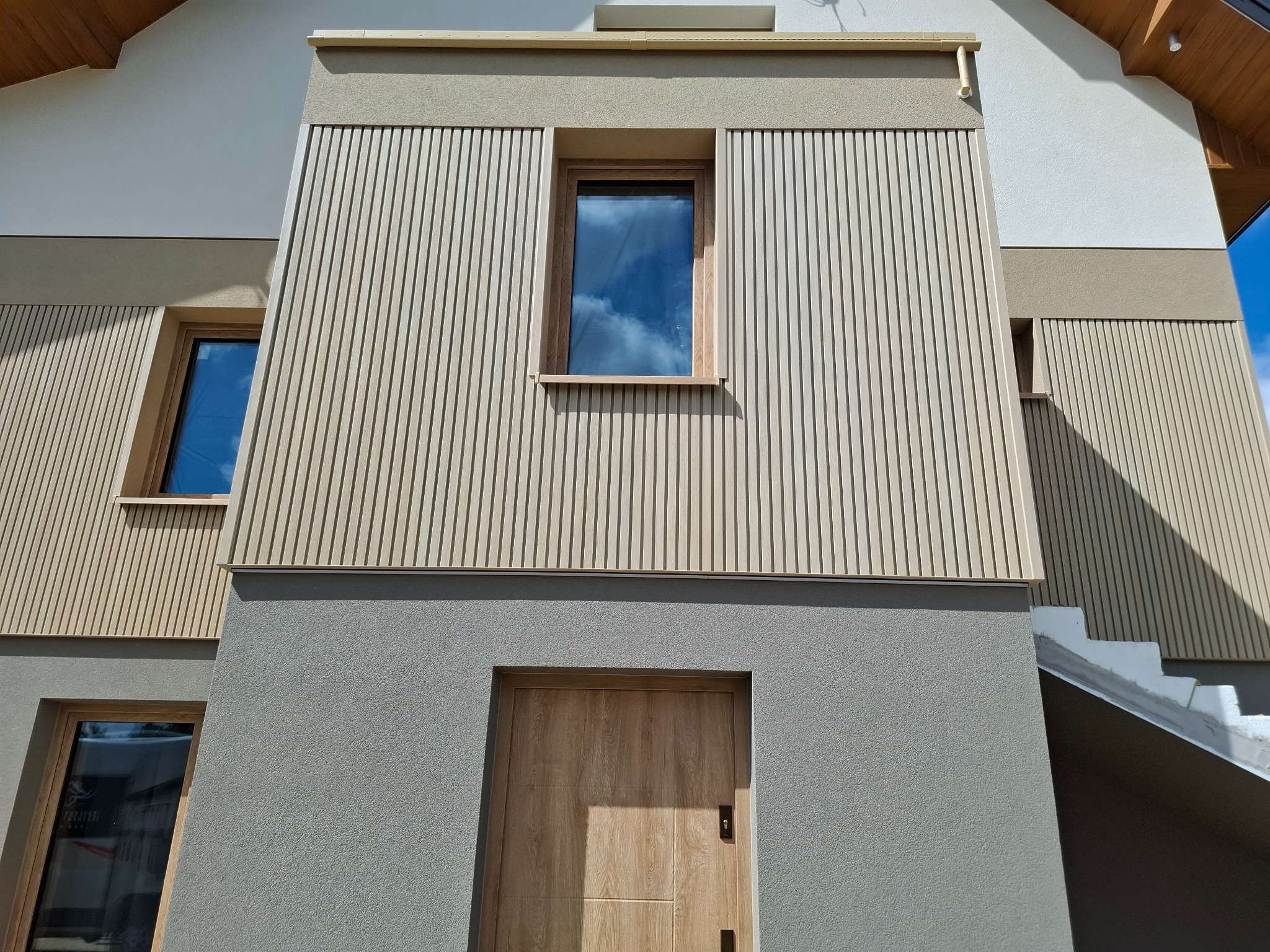 Nowoczesna elewacja domu z drewnianymi panelami pionowymi i szarym tynkiem. Widoczne okna z drewnianymi ramami i drzwi wejściowe w podobnym stylu.