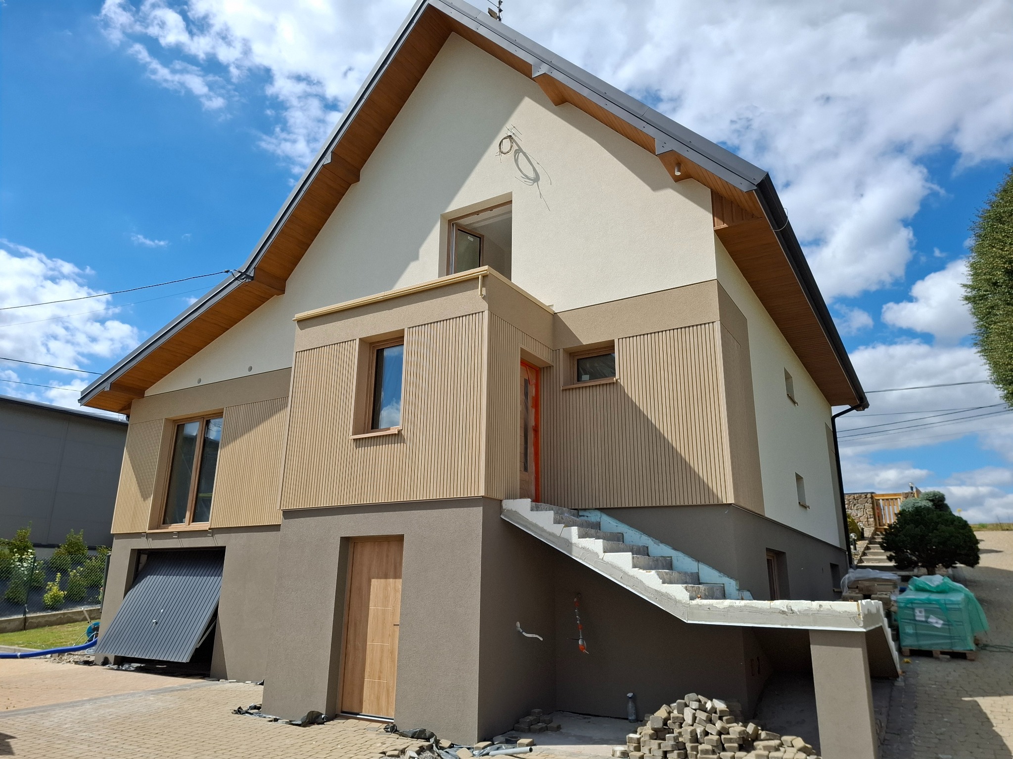 Dwukondygnacyjny dom z elewacją w odcieniach beżu i brązu, z częściowo otwartą bramą garażową i schodami wejściowymi w trakcie wykończenia, na tle błękitnego nieba.