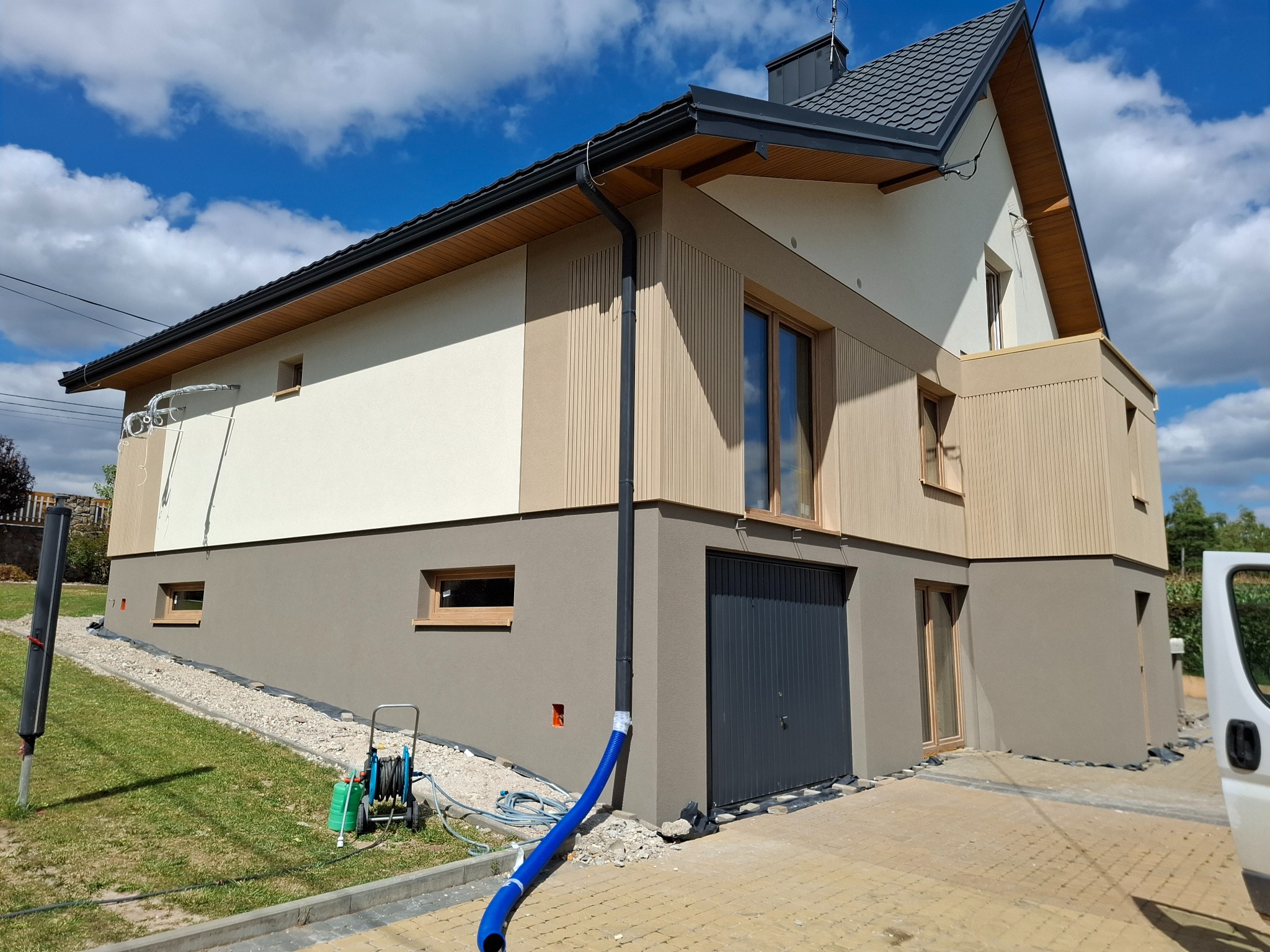 Nowoczesny dom po renowacji elewacji: połączenie beżu, brązu i pionowych paneli. Widoczne narzędzia i niebieski wąż sugerują trwające prace wykończeniowe.