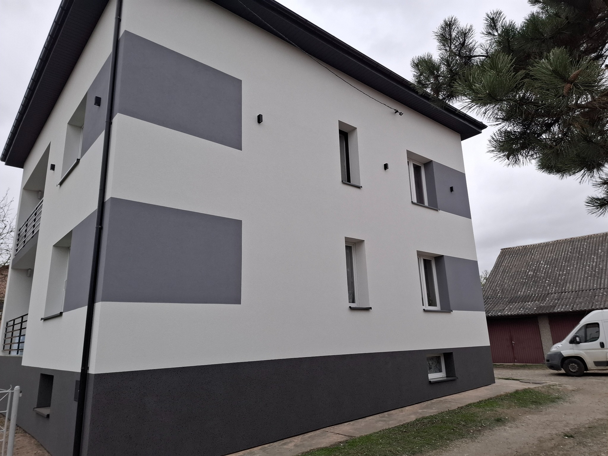 Dwukondygnacyjny budynek z elewacją w odcieniach szarości i bieli, z ciemnoszarą dolną częścią i pasami. Widoczne okna i fragment dachu. Obok drzewo i budynek gospodarczy.