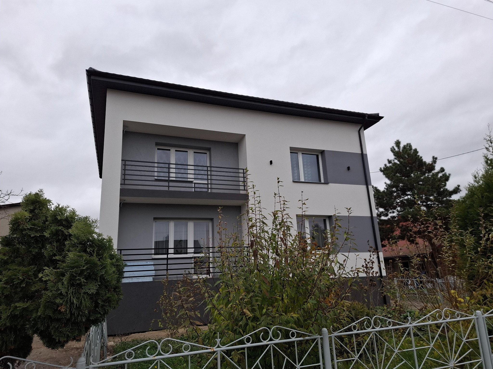 Dwupiętrowy dom z balkonami, świeżo po remoncie elewacji w odcieniach bieli i szarości, widoczny zza metalowego ogrodzenia i zieleni. Pochmurne niebo w tle.
