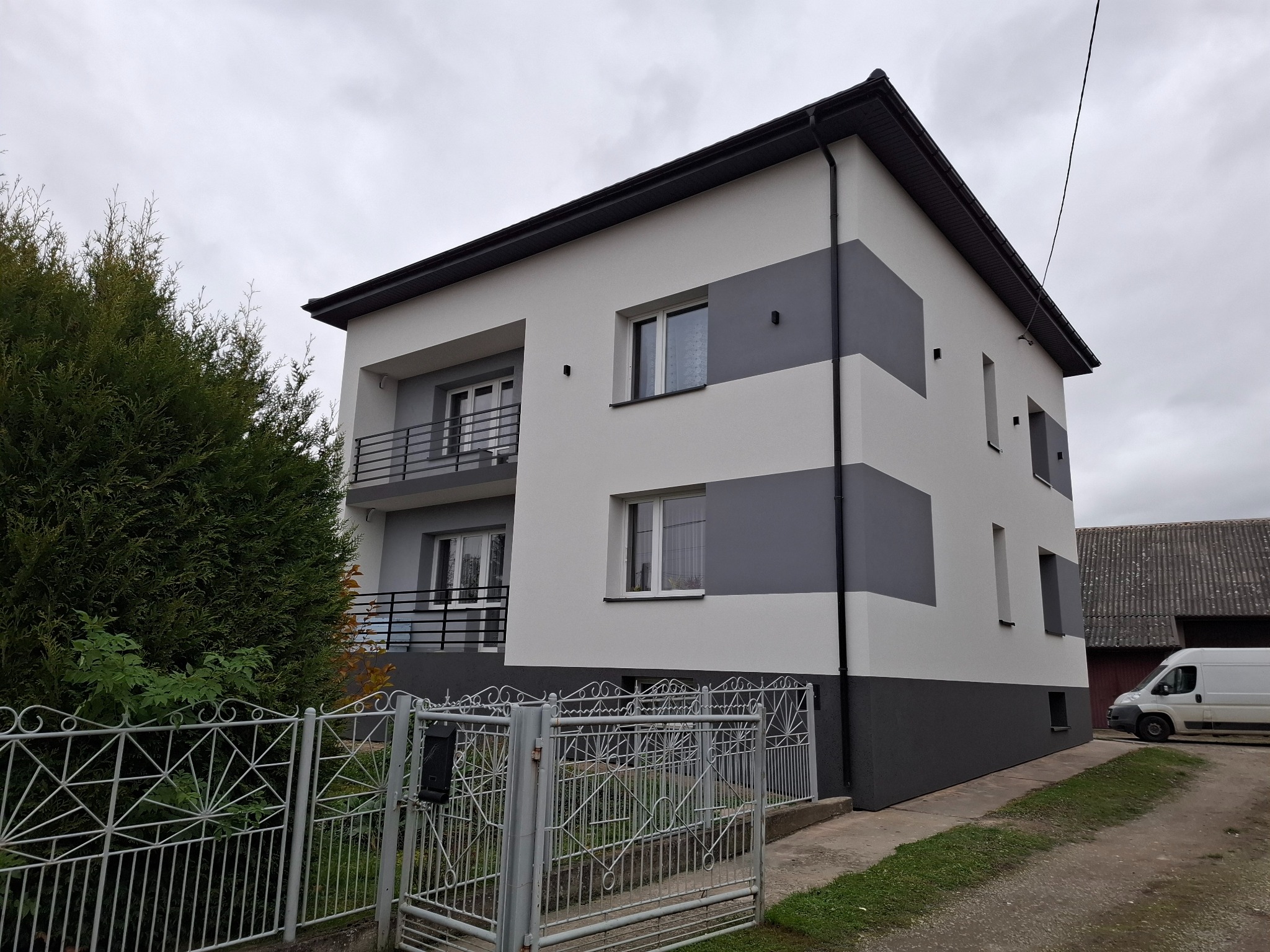 Dwukondygnacyjny dom z elewacją w odcieniach szarości i bieli, widoczny zza metalowego ogrodzenia. Na podwórku zaparkowany biały bus.