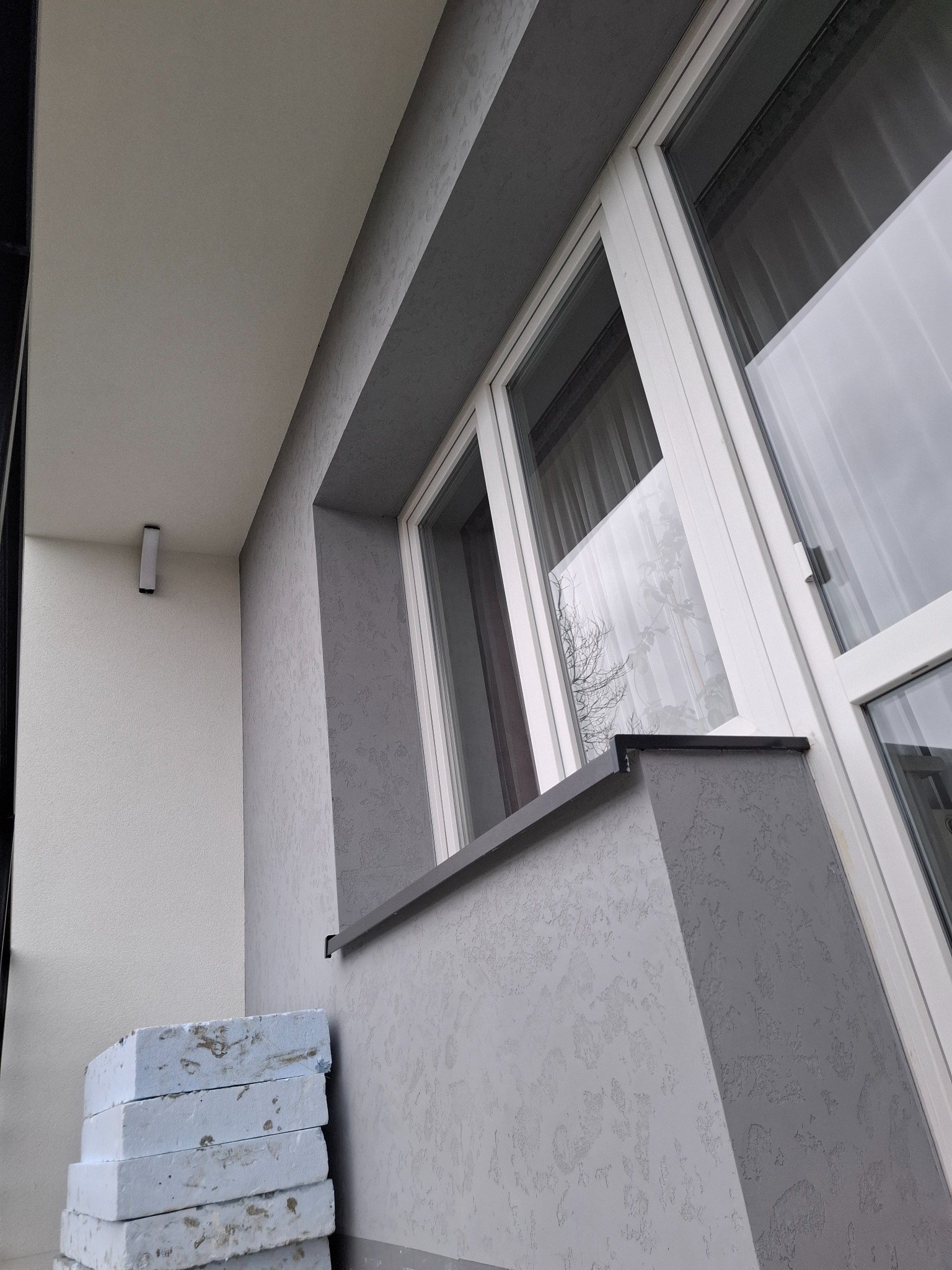 Szara elewacja balkonu z oknem PCV, widoczny fragment ściany z fakturą i stos styropianu. Ujęcie z dołu, podkreślające detale wykończenia.
