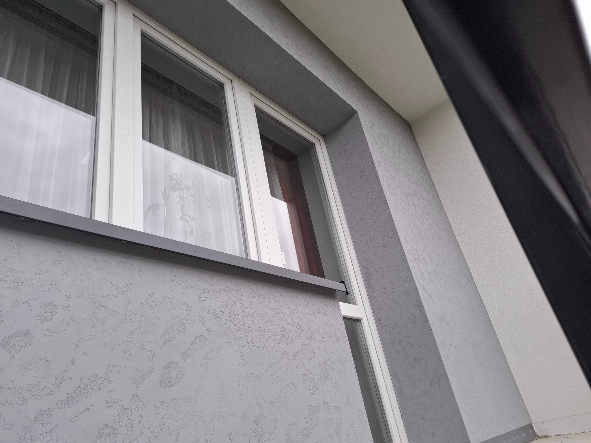 Szara elewacja z oknem PCV, widok z dołu. Dekoracyjna faktura tynku, białe ramy okienne. Ujęcie z niskiej perspektywy podkreśla strukturę elewacji.