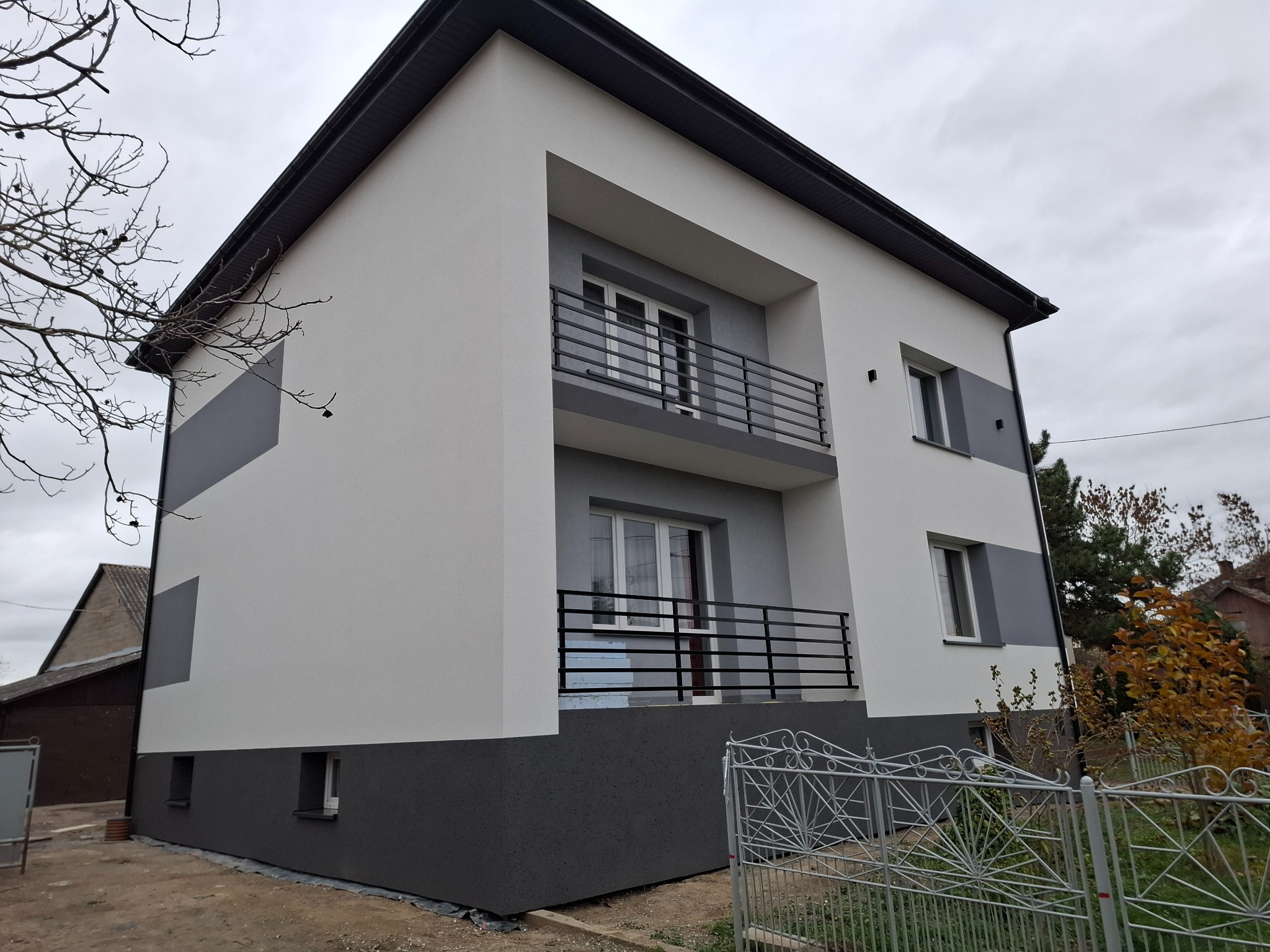 Dwukondygnacyjny dom z ocieploną elewacją w odcieniach szarości i bieli, z balkonami z czarną balustradą, widok z boku na tle pochmurnego nieba.
