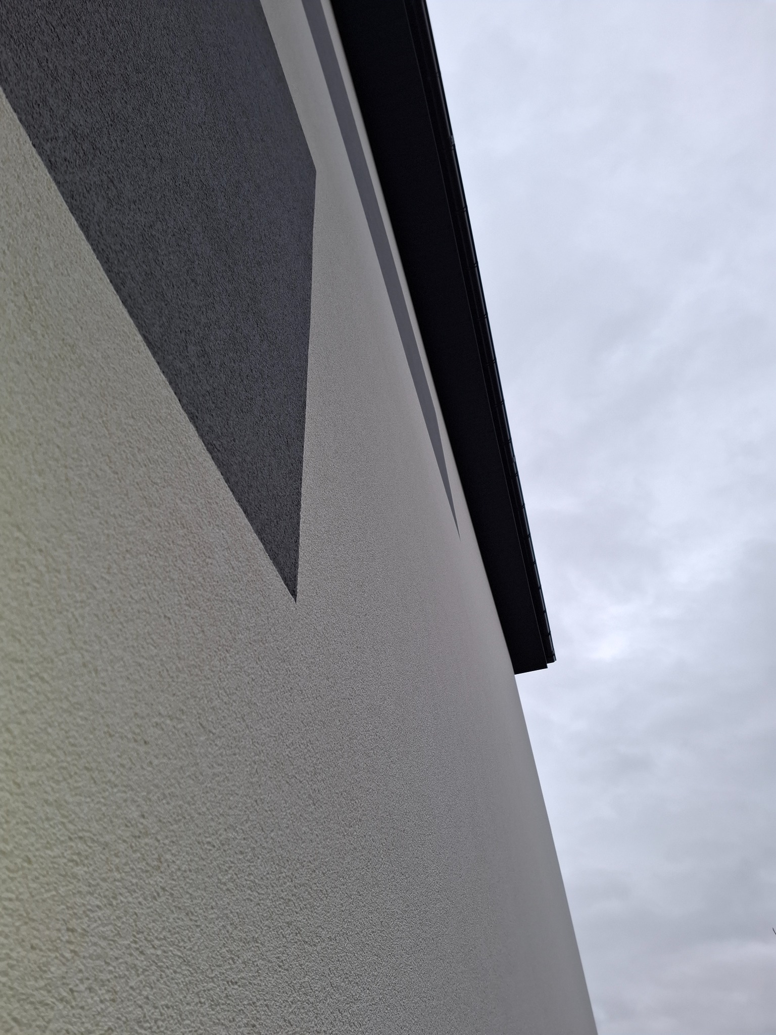 Fragment elewacji budynku z dekoracyjnym, geometrycznym wzorem w odcieniach szarości i beżu, widoczne orynnowanie na tle pochmurnego nieba.