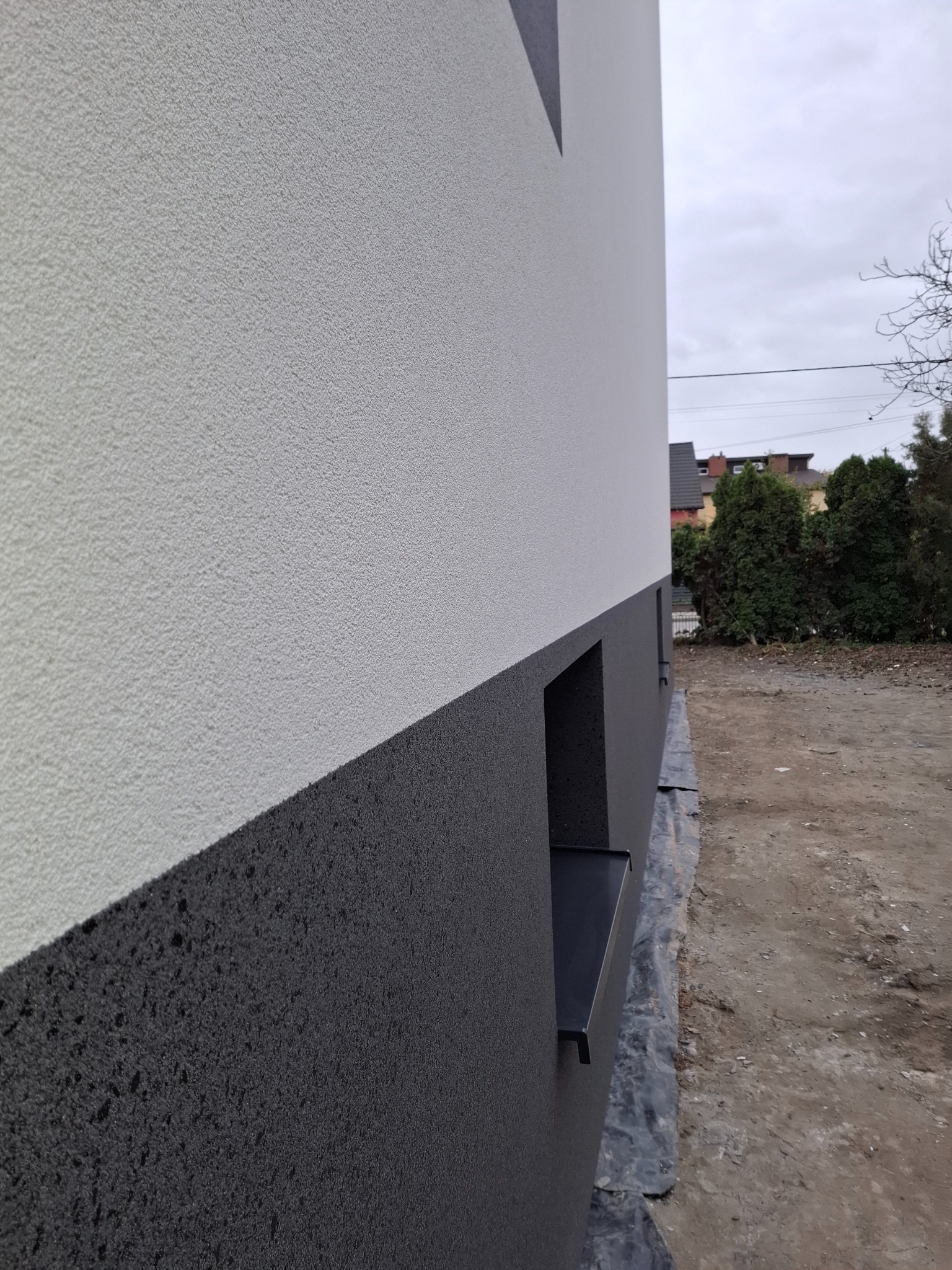 Dwukolorowa elewacja budynku z wyraźną fakturą tynku, widok z boku, z fragmentem okna i zabezpieczeniem gruntu folią. Szara, pochmurna pogoda w tle.