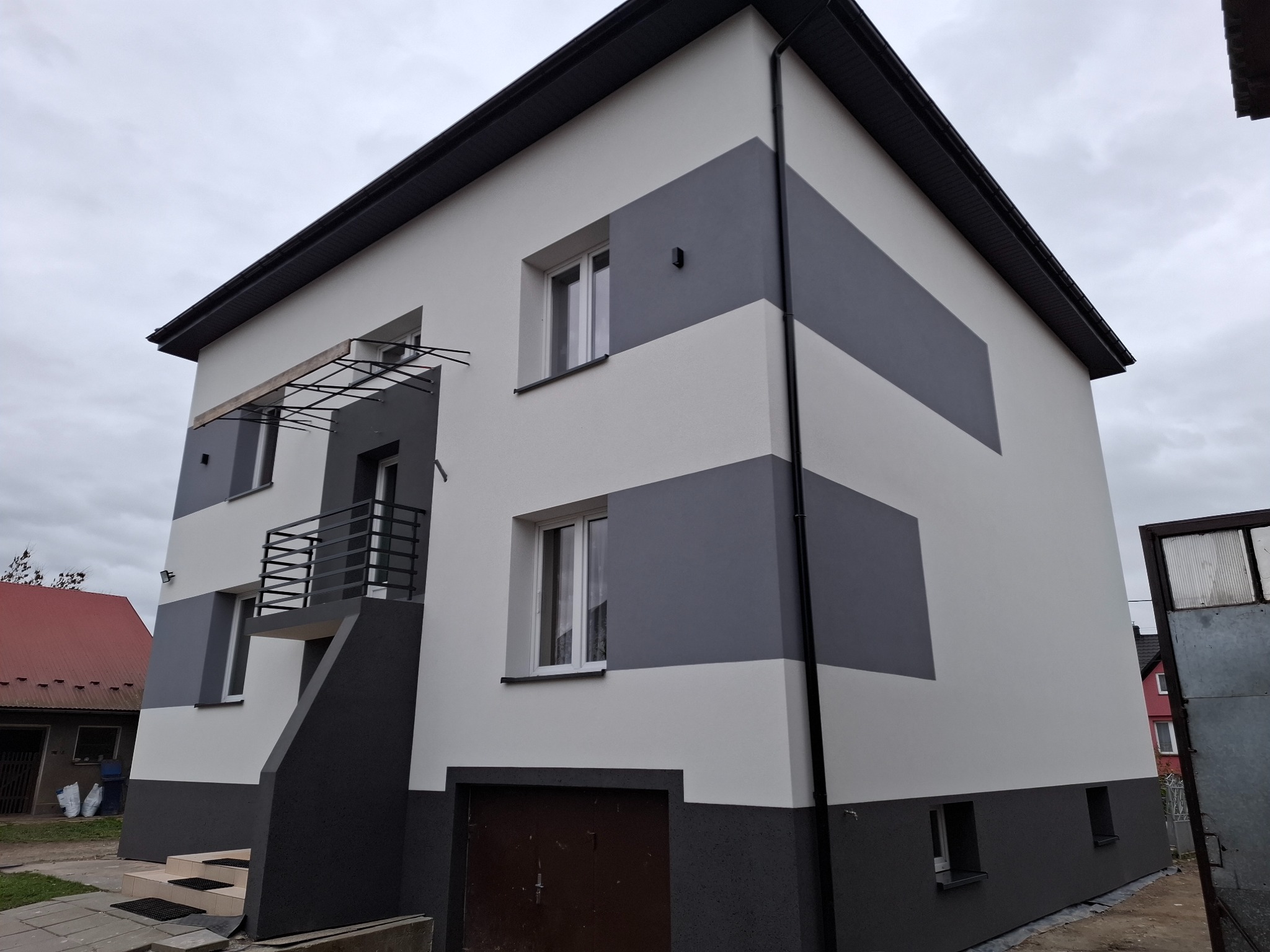 Dwukondygnacyjny dom z elewacją w odcieniach szarości i bieli, z balkonem i garażem. Widoczne detale wykończenia, takie jak rynny i parapety. Pochmurne niebo w tle.