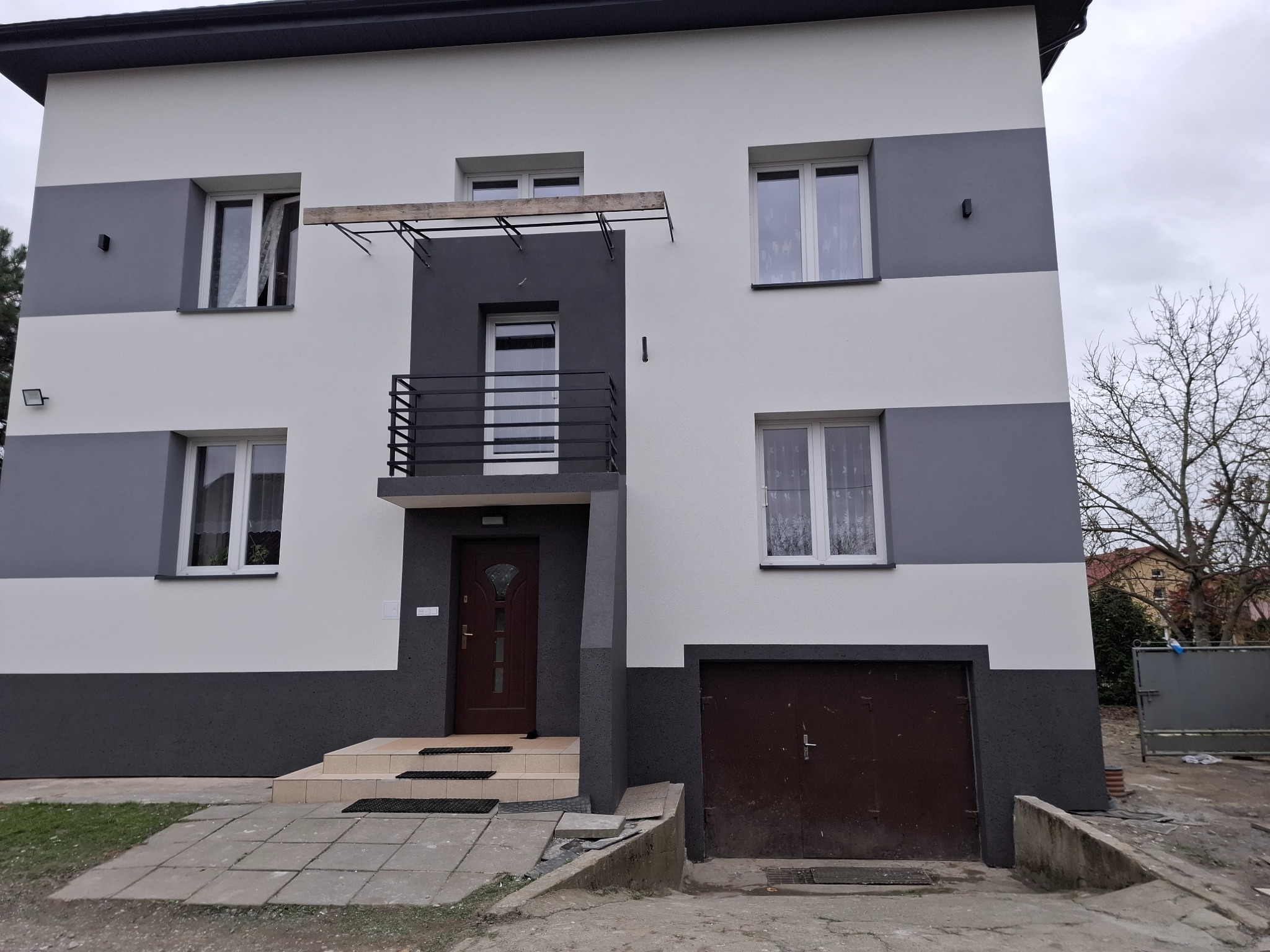 Dwukondygnacyjny dom po renowacji elewacji w odcieniach szarości i bieli, z ciemnobrązowymi drzwiami wejściowymi i garażowymi. Widoczne detale wykończenia.