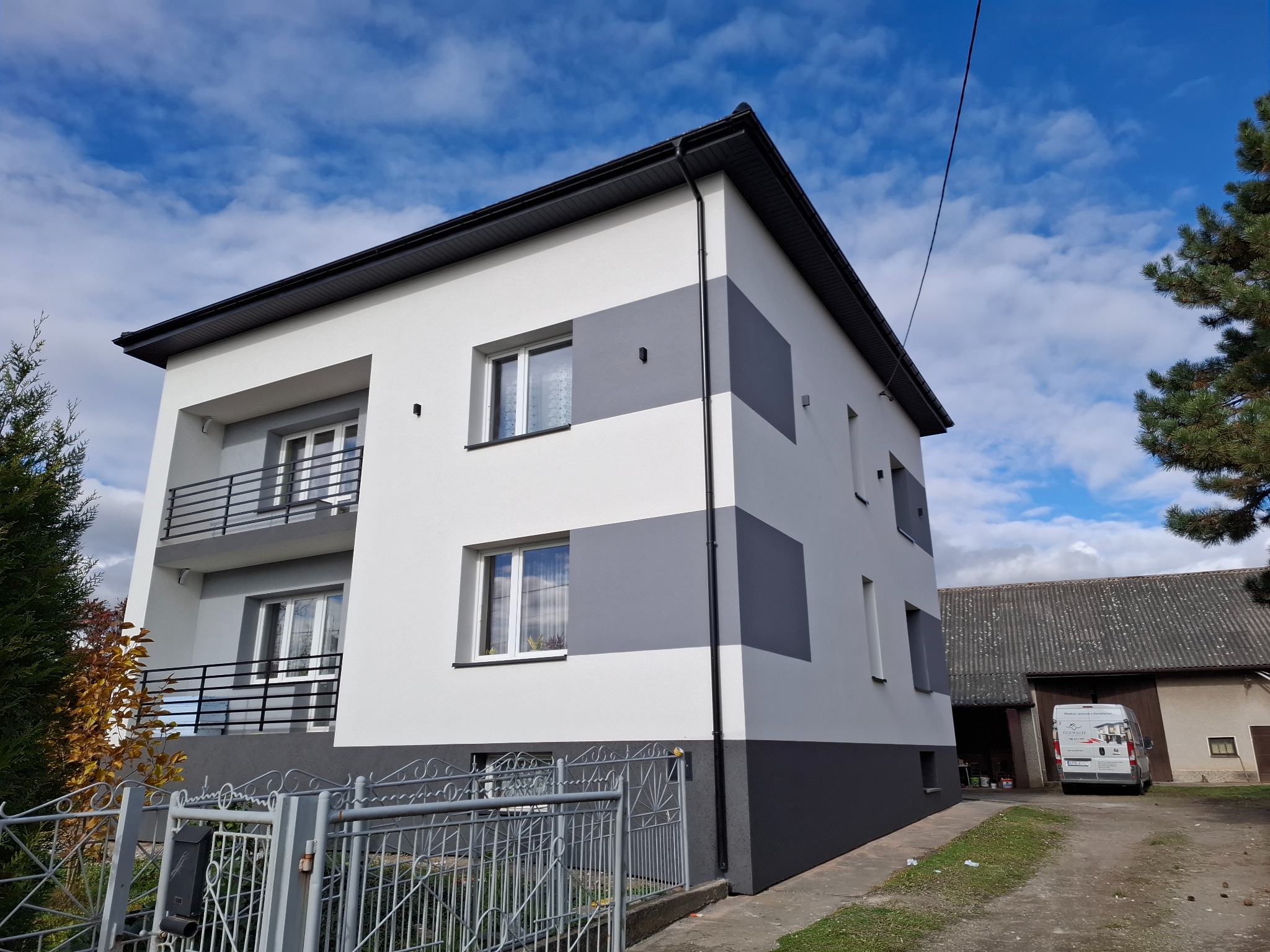 Dwukondygnacyjny dom po remoncie elewacji w odcieniach szarości i bieli, z balkonami i szarą metalową bramą. Na podwórku zaparkowany biały bus.