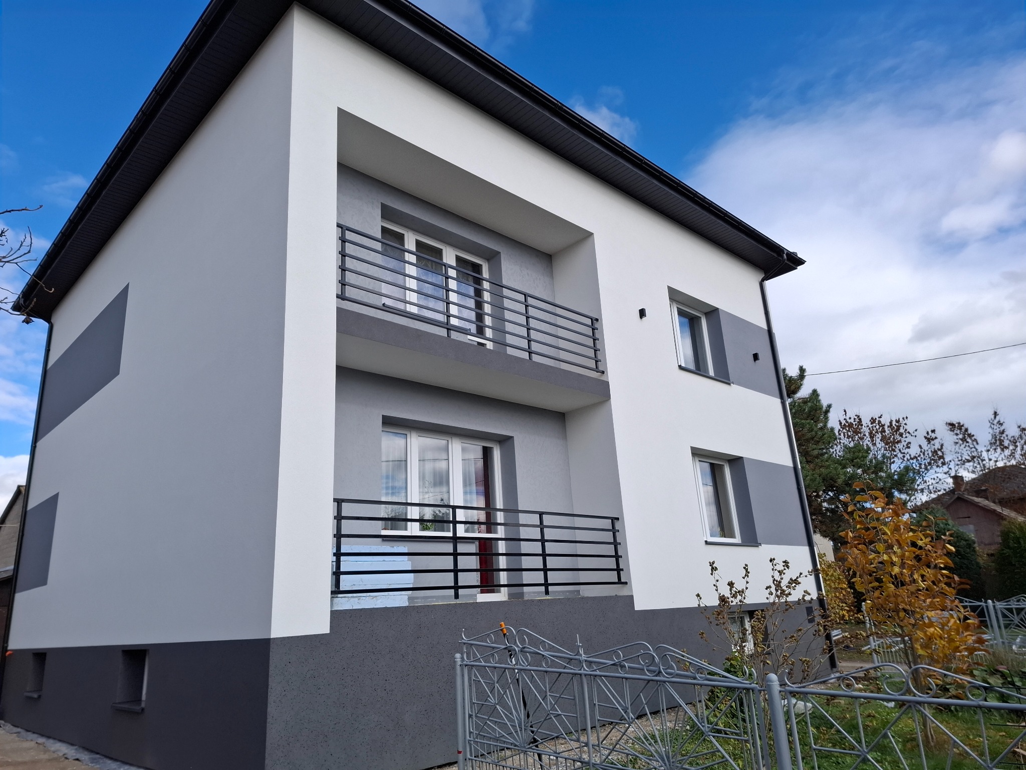 Dwupiętrowy dom z balkonami, świeżo po remoncie elewacji w odcieniach szarości i bieli, z czarnymi balustradami. Widok z dołu, błękitne niebo w tle.