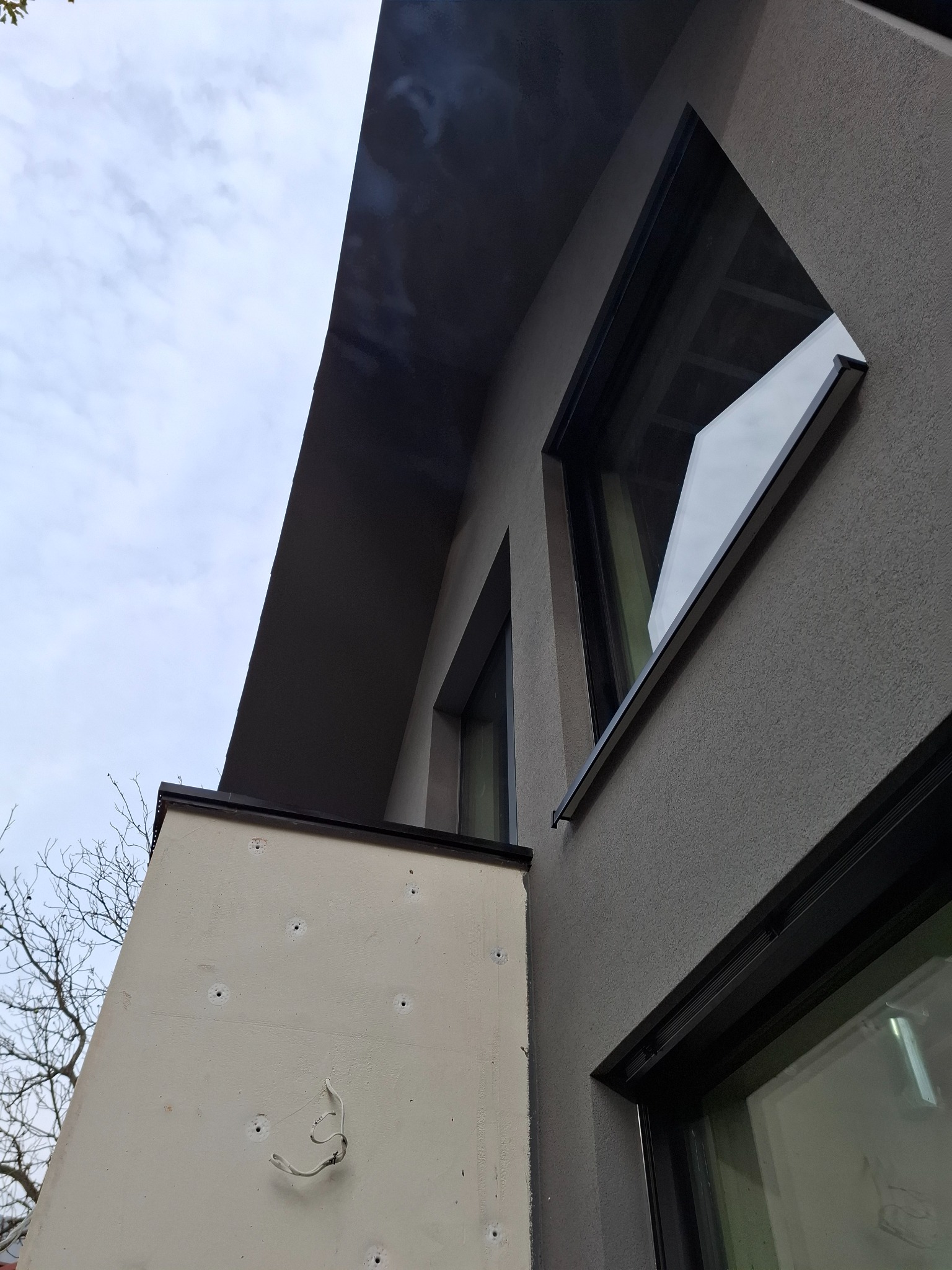 Szara elewacja budynku z geometrycznymi oknami i fragmentem ściany z widocznymi otworami, sfotografowana pod kątem z dołu w pochmurny dzień.