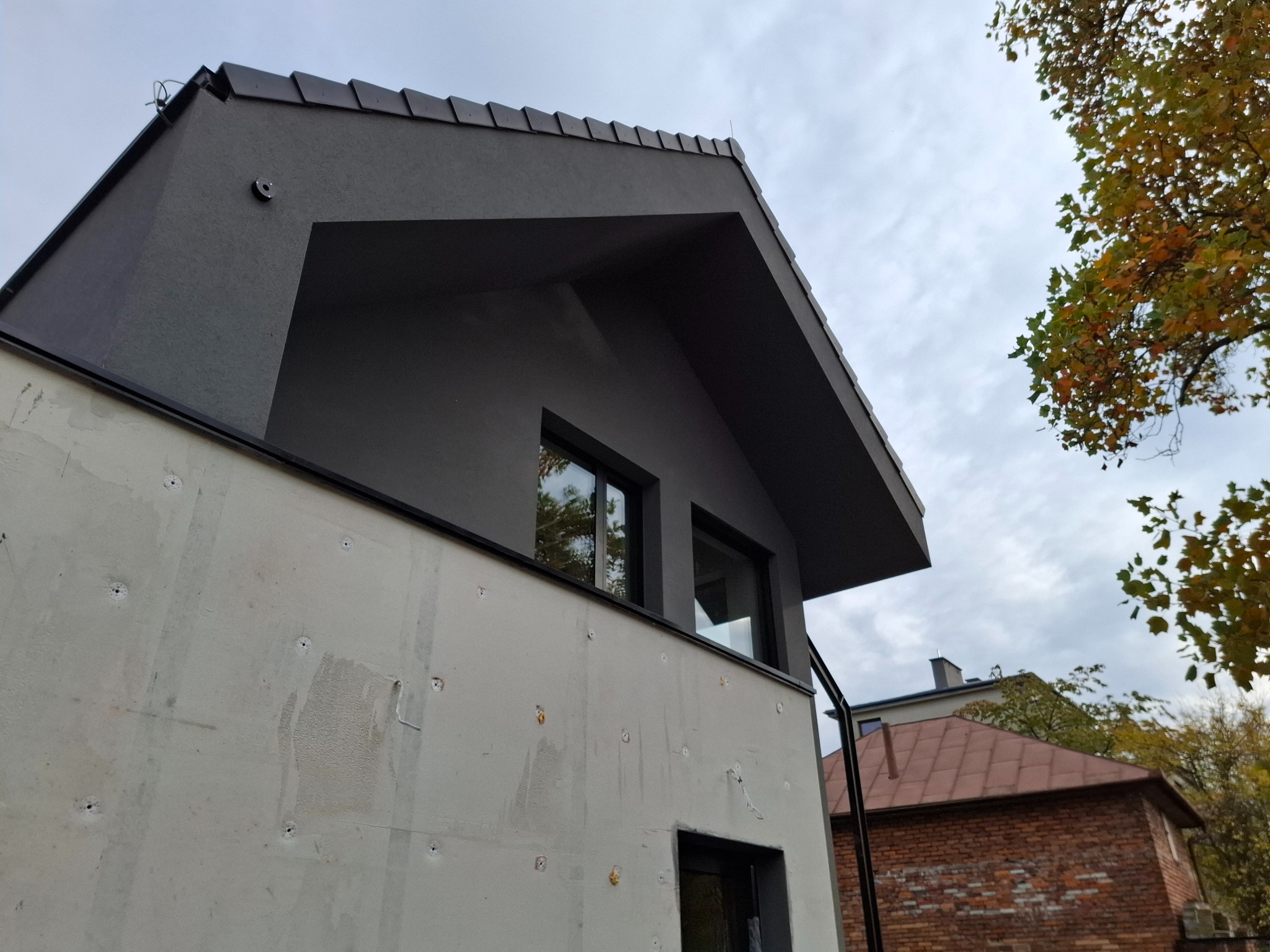 Szara elewacja domu w trakcie ocieplania, widoczne otwory montażowe i fragment dachu. Jesienne drzewo w tle.