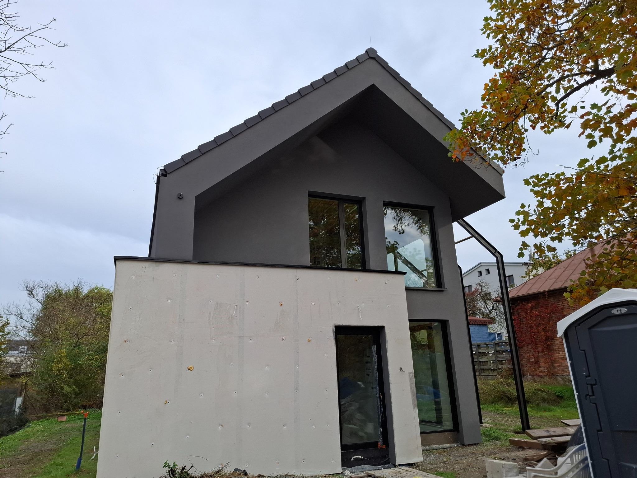 Nowoczesny dom jednorodzinny z szarą elewacją i fragmentem ściany w trakcie prac wykończeniowych. Widoczne okna i drzwi z czarnymi ramami, jesienne drzewo w tle.