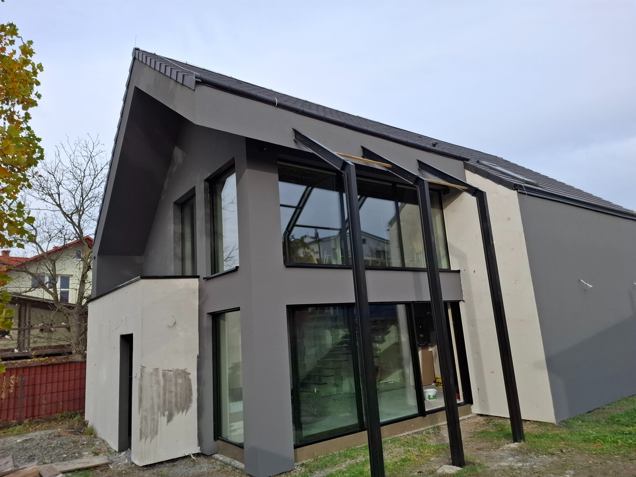 Nowoczesny dom z szarą elewacją i dużymi oknami, widok z zewnątrz. Część elewacji w jasnym odcieniu, prawdopodobnie w trakcie prac wykończeniowych. Widoczne drzewa w tle.