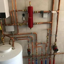 Kompleksowa instalacja grzewcza z miedzianymi rurami, czerwonym separatorem powietrza Flamco i białym zasobnikiem ciepłej wody na tle jasnych płytek ceramicznych.