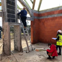 Budowa domu w trakcie: murarz na drabinie przy ścianie z czerwonej cegły, dwie osoby analizują plany, widoczne betonowe słupy i drewniana konstrukcja dachu.